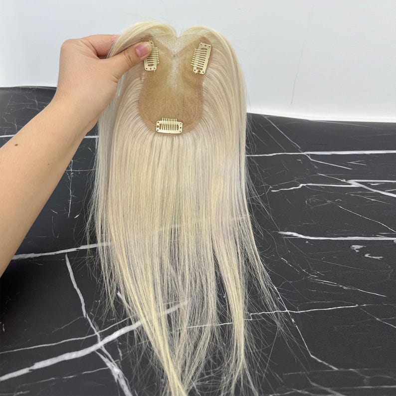 May include: A blonde hair topper with three small clips attached. The hairpiece is a light, natural blonde color and appears to be made of real hair. The clips are a light gold color.