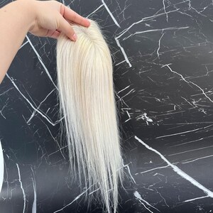 May include: A blonde hair topper with long, straight strands. The hairpiece is held up against a black and white marble background. The hair topper appears to be made of real hair.