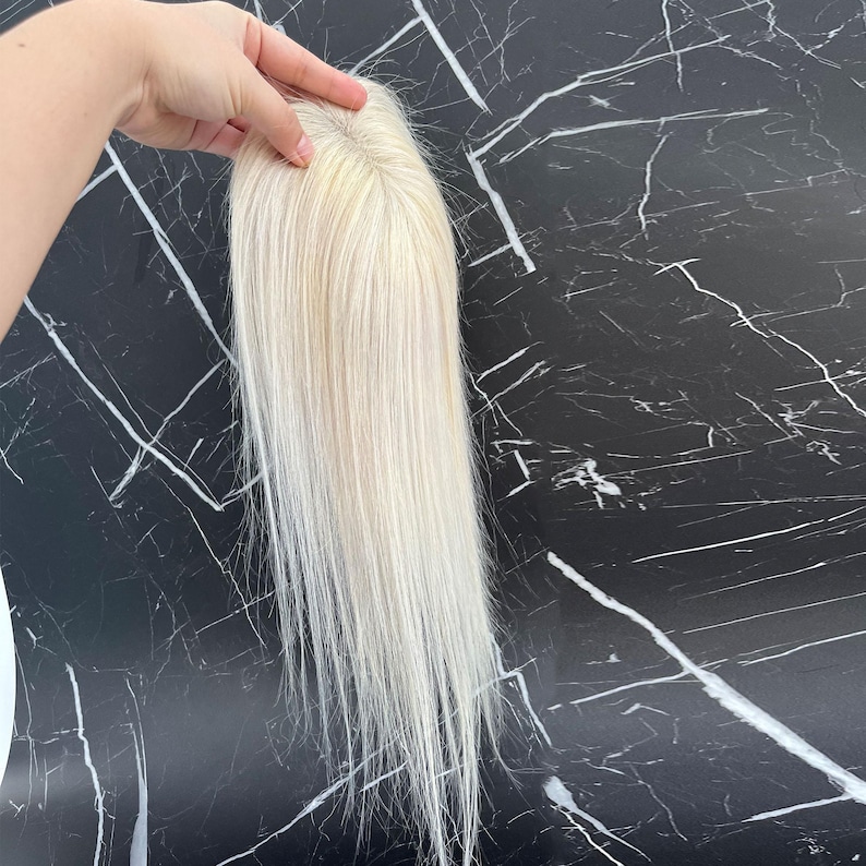 May include: A blonde hair topper with long, straight strands. The hairpiece is held up against a black and white marble background. The hair topper appears to be made of real hair.