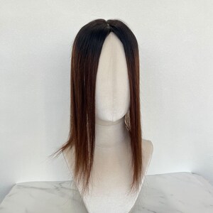 以下が含まれることがあります： 長くてストレートな髪の毛が付いた、茶色と黒のヘアトッパー。ヘアピースは、白い背景にマネキンの頭に表示されています。髪の根元は濃い茶色で、毛先に向かって明るい茶色に変化しています。