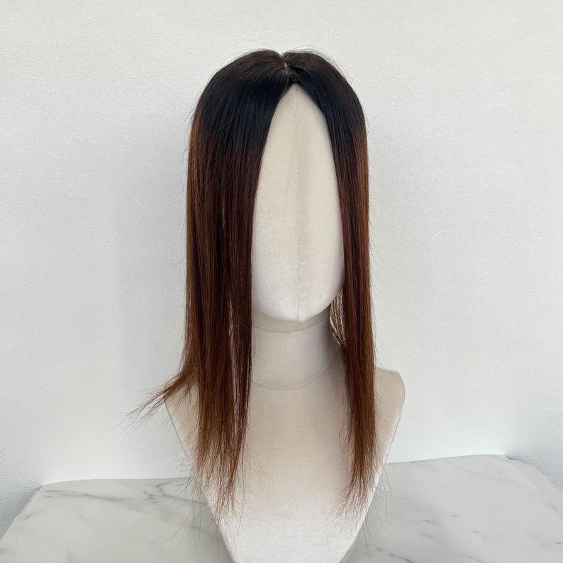 以下が含まれることがあります： 長くてストレートな髪の毛が付いた、茶色と黒のヘアトッパー。ヘアピースは、白い背景にマネキンの頭に表示されています。髪の根元は濃い茶色で、毛先に向かって明るい茶色に変化しています。