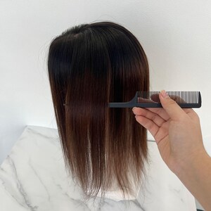 以下が含まれることがあります： 黒い櫛を使って、濃い茶色の髪をとかしています。髪はストレートで、根元から毛先にかけて色が薄くなる、わずかなオンブレ効果があります。櫛は手で持たれており、ヘアケアツールの使用方法を示しています。