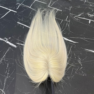 May include: A light blonde hair topper with a natural-looking part. The hairpiece is made of fine, straight hair and is designed to blend seamlessly with natural hair. The hairpiece is on a black comb.