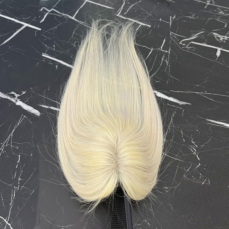 May include: A light blonde hair topper with a natural-looking part. The hairpiece is made of fine, straight hair and is designed to blend seamlessly with natural hair. The hairpiece is on a black comb.