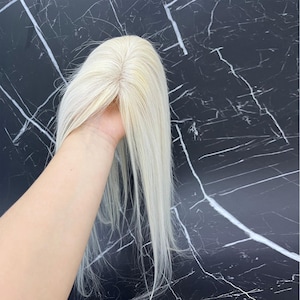 May include: A blonde hair topper with long, straight strands. The hairpiece is held up, showcasing the natural-looking part and the light color. The hairpiece appears to be made of human hair.