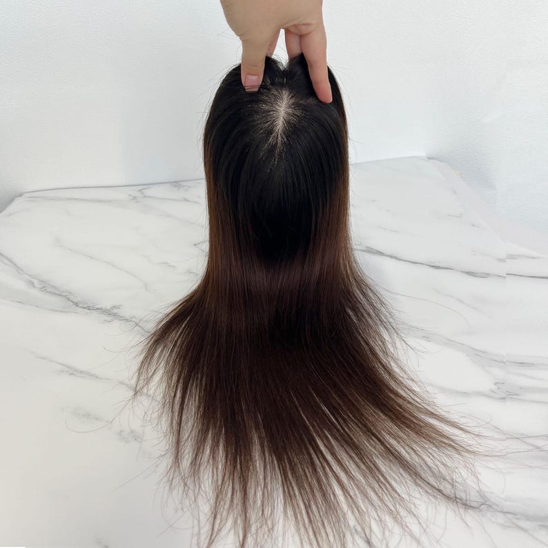 以下が含まれることがあります： 自然なヘアラインのダークブラウンのヘアトッパー。髪は長くストレートで、わずかにオンブル効果があります。ヘアピースは白い背景に対して手で持たれています。