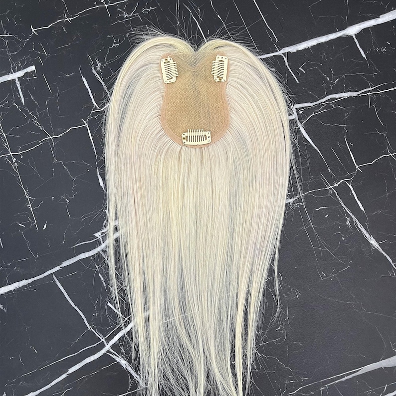 May include: A light blonde hair topper with three small clips attached to a beige base. The hair is straight and falls down from the base. The background is a black and white marble pattern.