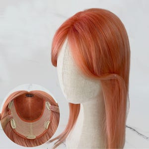 Könnte beinhalten: Ein pfirsichfarbener Haarteil mit langem, glattem Haar und Pony. Das Haarteil wird auf einem Schaufensterpuppenkopf präsentiert. Das Haarteil hat Clips zur Befestigung und eine Netzbasis.