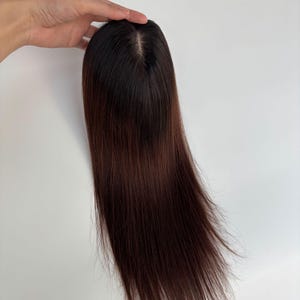 以下が含まれることがあります： 長いストレートヘアのダークブラウンのヘアピース。ヘアピースは頭頂部に自然な分け目があり、白い背景に対して手で持たれています。髪の色は根元が濃く、明るい茶色に変化します。