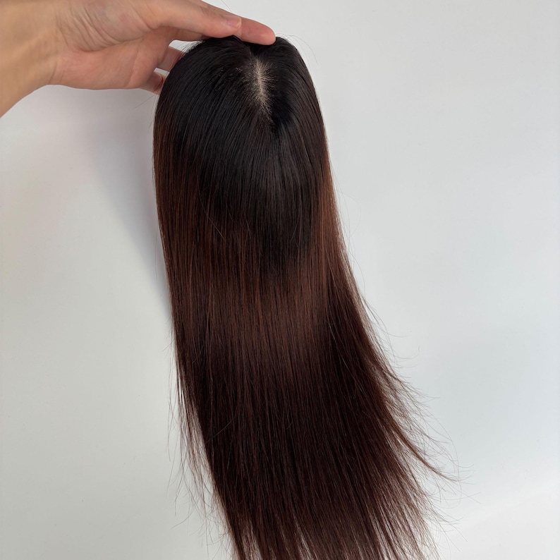 以下が含まれることがあります： 長いストレートヘアのダークブラウンのヘアピース。ヘアピースは頭頂部に自然な分け目があり、白い背景に対して手で持たれています。髪の色は根元が濃く、明るい茶色に変化します。