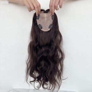 Può includere: Un'extension per capelli castano scuro con una texture ondulata. L'extension ha una piccola base color carne con clip per il fissaggio. I capelli sono tenuti da due mani su uno sfondo bianco.