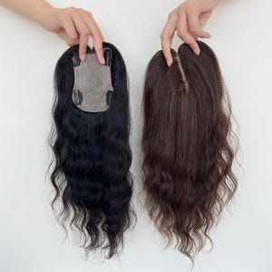 May include: Two hair toppers are displayed against a white background. One is black with wavy texture, and the other is brown with a similar wavy style. Both have a mesh base with clips for attachment, showcasing hair volume and color options.