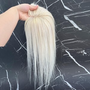 May include: A blonde hair topper with long, straight strands. The hairpiece has a light, natural color with subtle highlights. The base of the topper is visible, showing the construction. The hair is held up against a black and white marble background.