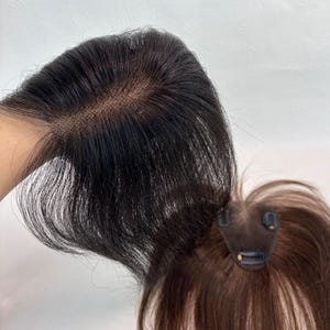 May include: Two hair toppers are displayed, one in dark brown and the other in black. The black hair topper is held up, showcasing the hair's natural-looking hairline. The brown hair topper has a clip attachment. Both toppers appear to be made of human hair.