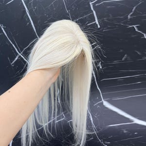 May include: A light blonde hair topper with long, straight strands. The hairpiece is held up against a black and white marble background. The hair topper appears to be made of human hair.