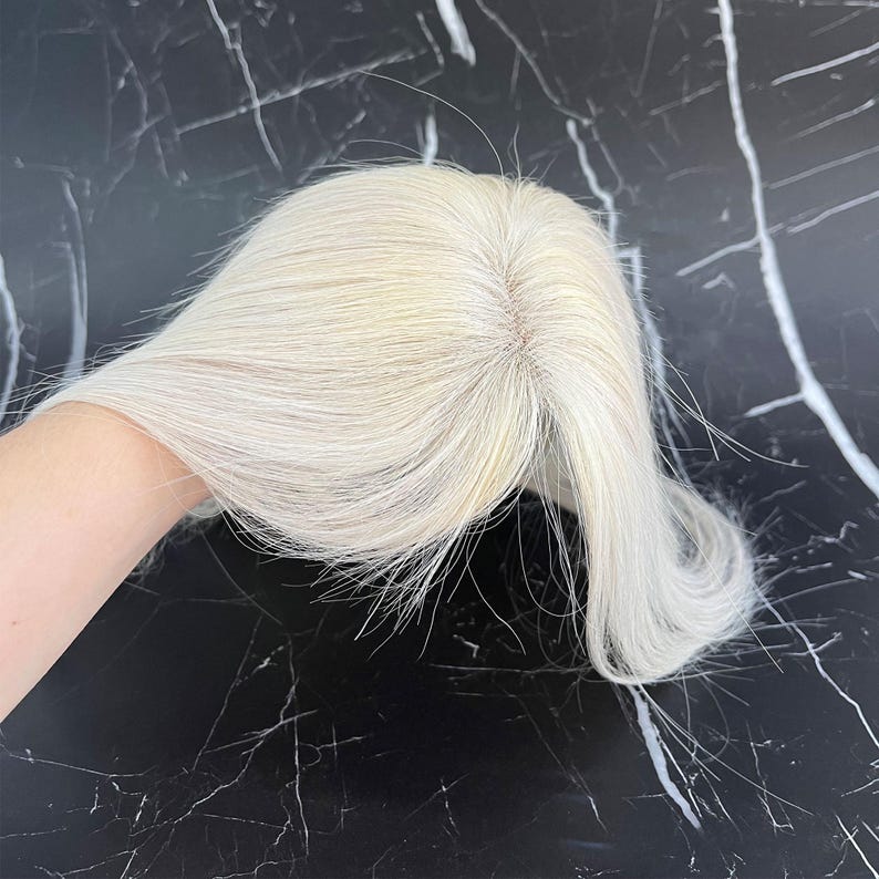 May include: A light blonde hair topper with a natural-looking part. The hair is straight and appears to be made of human hair. The topper is designed to add volume and coverage to the top of the head.