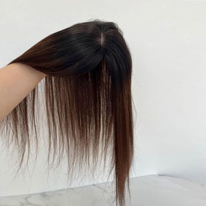 以下が含まれることがあります： 黒い根元の部分がある、濃い茶色のヘアートッパー。髪はストレートで長く、自然な質感です。ヘアピースは白い背景に対して保持されています。