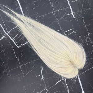 May include: A light blonde hair topper, likely human hair, with a natural-looking part. The hairpiece is laid flat against a black and white marble surface, showcasing its color and texture. The hair is finely textured and appears to be styled.