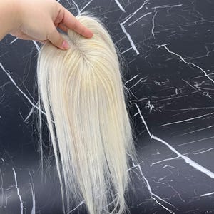 May include: A blonde hair topper with long, straight strands. The hairpiece is held by a hand, showcasing the natural-looking roots and the overall color. The background is a black surface with white veining.