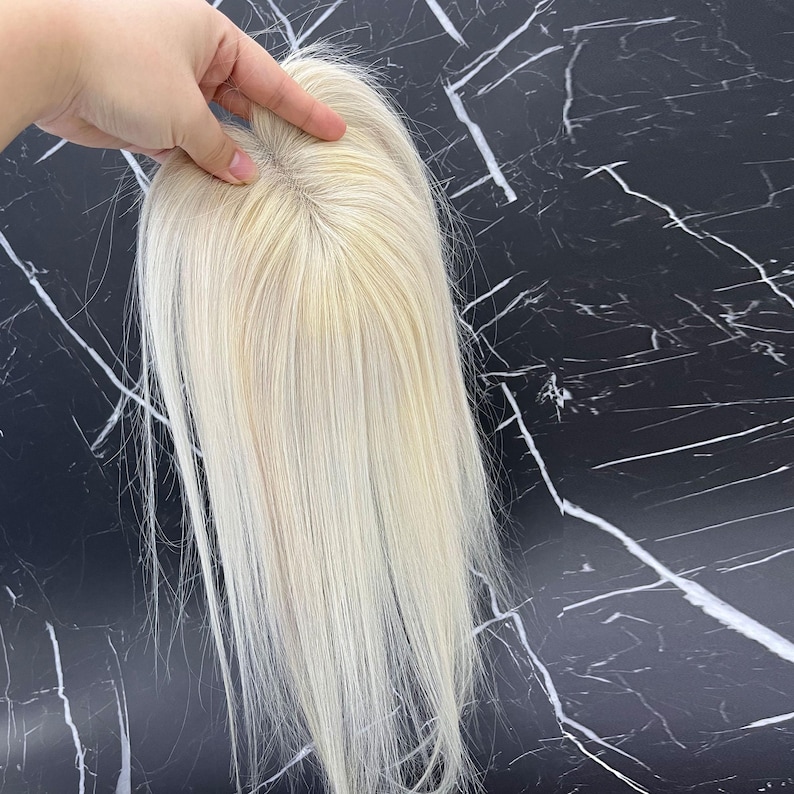 May include: A blonde hair topper with long, straight strands. The hairpiece is held by a hand, showcasing the natural-looking roots and the overall color. The background is a black surface with white veining.
