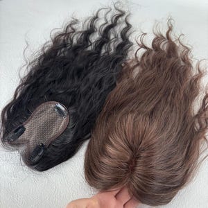 Op de afbeelding: Twee haartoppers met golvend haar. Eén is zwart en de andere is bruin. Elke topper heeft een mesh basis met clips voor bevestiging. Het haar is gestyled in losse golven en biedt een natuurlijke uitstraling. Deze haarstukken zijn ontworpen om volume en dekking toe te voegen.