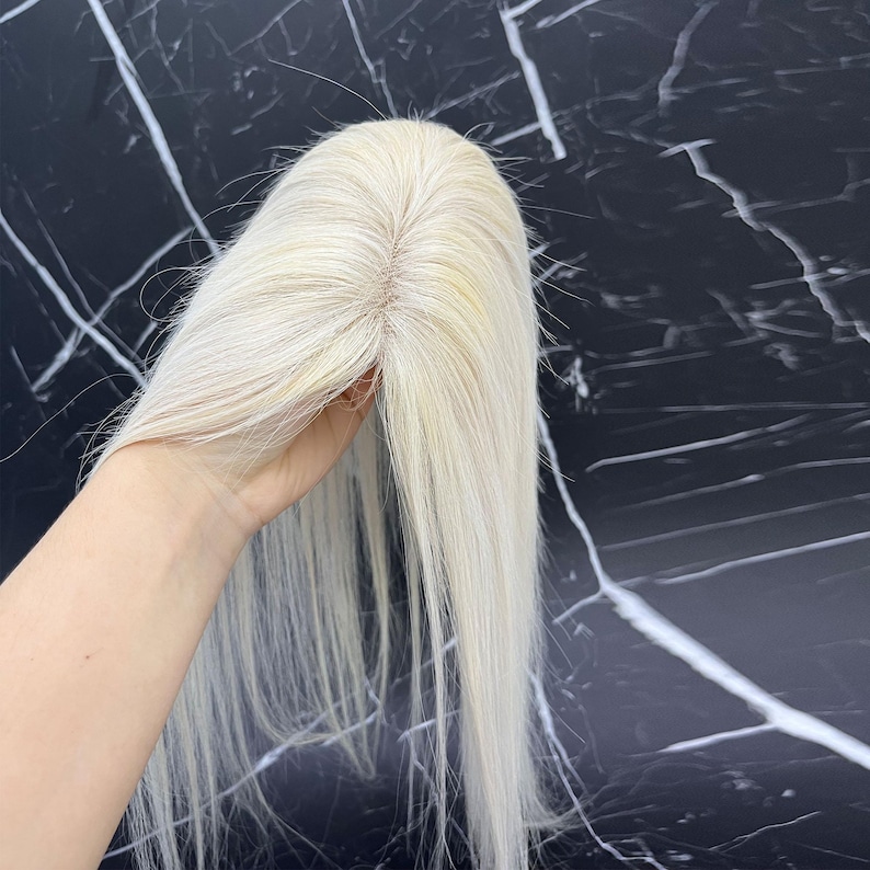 May include: A light blonde hair topper with long, straight strands. The hairpiece is held up against a black and white marble background. The hair topper appears to be made of human hair.