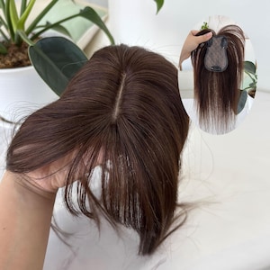 Pode incluir: Uma touca de cabelo castanho com uma risca de aspeto natural. A peça de cabelo é segurada, mostrando a base e o comprimento do cabelo. O cabelo é de uma cor castanha escura rica, e o estilo parece ser liso.