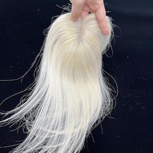 May include: A light blonde hair topper with long, straight strands. The hairpiece is held up against a dark background, showcasing the natural color and texture of the hair. The topper appears to be made of human hair.
