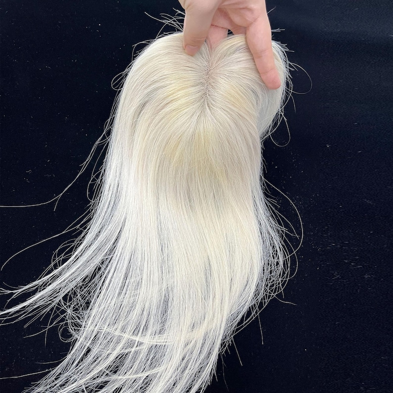 May include: A light blonde hair topper with long, straight strands. The hairpiece is held up against a dark background, showcasing the natural color and texture of the hair. The topper appears to be made of human hair.