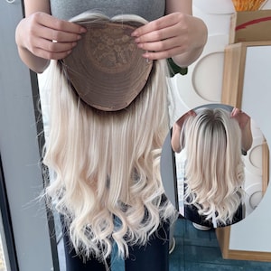 Puede incluir: Una peluca rubia con cabello largo y ondulado. Se ve la gorra de la peluca, que muestra una base marrón con un diseño de encaje. El cabello es rubio claro con raíces más oscuras, y el estilo es largo y rizado.