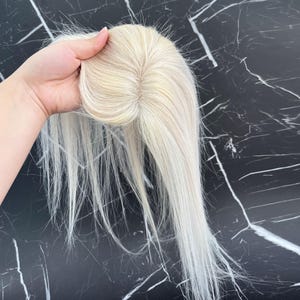 May include: A blonde hair topper with long, straight strands. The hairpiece is held up, showcasing the base and the natural-looking hair color. The hairpiece appears to be made of high-quality synthetic or human hair.