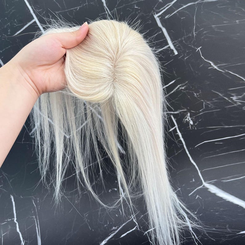 May include: A blonde hair topper with long, straight strands. The hairpiece is held up, showcasing the base and the natural-looking hair color. The hairpiece appears to be made of high-quality synthetic or human hair.