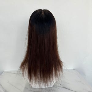 以下が含まれることがあります： ダークブラウンからブラックのグラデーション効果のある、長くてストレートなウィッグ。髪は自然な分け目でスタイリングされ、肩まで届きます。ウィッグは白い背景に白いマネキンの頭に展示されています。