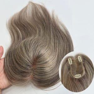 以下が含まれることがあります： アッシュブラウンのストレートヘアに繊細なハイライトが入ったヘアピース。ベース部分には小さなメッシュ状のセンターパーツがあり、裏側には髪にしっかりと固定するための3つの金属製クリップが取り付けられています。