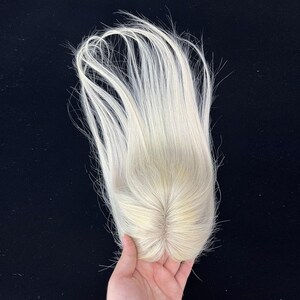 May include: A light blonde hair topper with long, straight strands. The hairpiece is held against a dark background, showcasing the natural color and texture of the hair. The topper appears to be a hair replacement system.
