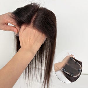Puede incluir: Una pieza de cabello castaño oscuro con una base de encaje, sostenida por las manos de una persona. El cabello es liso y largo, con una raya de aspecto natural. La base de encaje tiene clips para una fijación segura.