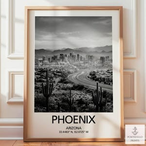 May include: Black and white framed print of Phoenix, Arizona. The image features a cityscape with a winding road, desert landscape, and mountains in the background. The word "PHOENIX" is printed at the bottom, along with the city's coordinates.