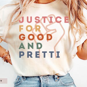 May include: Ivory-coloured t-shirt featuring the words "JUSTICE FOR GOOD AND PRETTI" in a colourful, stacked design. A raised fist graphic is behind the text. The shirt is a casual style.