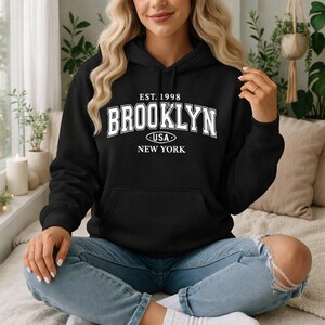 May include: Black hoodie featuring the word "BROOKLYN" in white block letters. The text "EST. 1998", "USA", and "NEW YORK" are also printed on the front. The hoodie has a front pocket and a drawstring hood.
