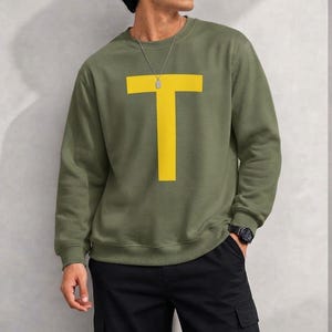May include: An olive green long-sleeve crewneck sweatshirt featuring a large, bold, yellow letter T printed on the center of the chest. The garment has ribbed cuffs and a ribbed neckline, providing a clean and structured look for casual wear.