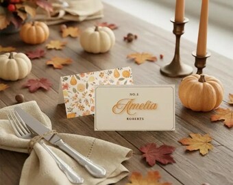 May include: A seasonal table setting featuring pumpkins, autumn leaves, and place cards. The place card reads "No. 5 Amelia Roberts". Cutlery is tied with twine on a linen napkin. Candles and a candlestick are also present.