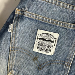 Peut inclure: Jean en denim bleu avec une étiquette rectangulaire blanche sur la poche arrière portant le texte noir Levi Strauss & Co. San Francisco Cal. Clothing. Une petite étiquette blanche avec le mot Levi's en noir est fixée sur le côté de la poche.