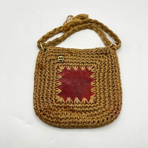Può includere: Una borsa a tracolla marrone all'uncinetto con una toppa quadrata in pelle rossa al centro. La borsa ha una tracolla intrecciata e un piccolo emblema color oro. La borsa è realizzata in un materiale di fibra naturale.