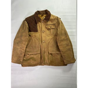 Puede incluir: Chaqueta de caza de color beige con cuello de pana marrón y parche acolchado en el hombro. La chaqueta tiene cuatro bolsillos delanteros, cierre de botones y un aspecto desgastado. Estilo vintage.