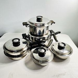 May include: A set of stainless steel cookware including a large stockpot, a medium saucepan, and two smaller pans, all featuring polished silver surfaces, black plastic handles, and matching lids with black circular knobs on top.