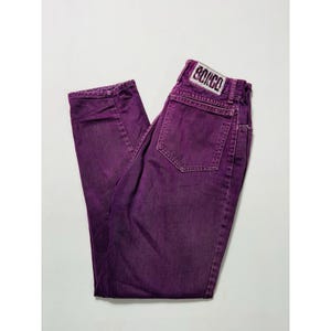 Puede incluir: Un par de vaqueros Bongo morados. Los vaqueros son de color morado liso y vibrante con un diseño clásico de cinco bolsillos. La etiqueta Bongo es visible en la cintura trasera. Los vaqueros están doblados y colocados sobre una superficie blanca.
