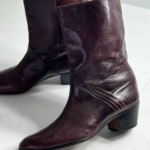 Puede incluir: Botas de cuero burdeos con punta afilada y tacón bajo. Las botas tienen un panel lateral decorativo con un emblema y detalles cosidos. El cuero tiene un ligero brillo y las botas parecen ser vintage. Altura del tacón: aproximadamente 5 cm.