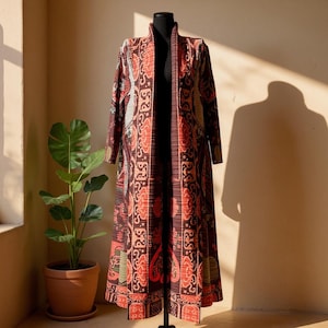 Può includere: Un cappotto lungo, aperto sul davanti, con un motivo intricato nei toni del rosso, marrone e arancione. Il cappotto è esposto su un manichino nero, mostrando la sua lunghezza e il suo design. Il motivo include elementi floreali e geometrici stilizzati.