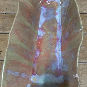 May include: A handmade ceramic serving dish with a unique, organic shape. The dish features a blend of warm colors, including peach, yellow, and red, with iridescent highlights. The interior has a rectangular shape with wavy edges.