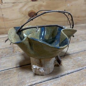 May include: A handmade ceramic bowl with a natural twig handle. The bowl has a speckled tan exterior with blue accents and sits atop a birch bark base. The rustic design is perfect for home decor.